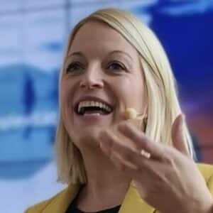 Frau mit blonden Haaren und Mikrofon spricht lächelnd, trägt einen gelben Blazer, Hintergrund unscharf.