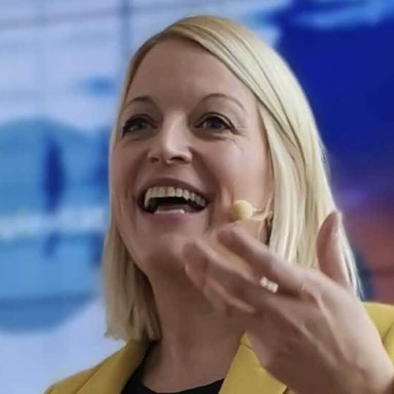 Frau mit blonden Haaren und Mikrofon spricht lächelnd, trägt einen gelben Blazer, Hintergrund unscharf.