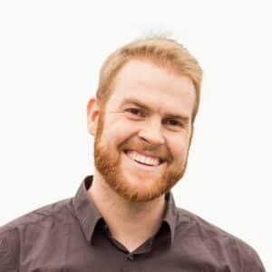 David Nitschke Lächelnder Mann mit blonder, kurzer Haarfrisur und Bart, trägt ein dunkles Hemd, vor hellem Hintergrund.