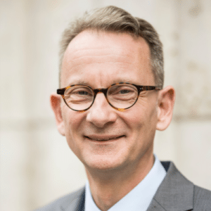 Porträt eines lächelnden Mannes mit Brille, grauem Anzug und hellblauer Krawatte, vor neutralem Hintergrund.