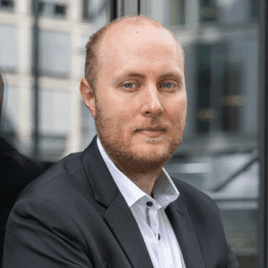 jan-schulte Mann mit kurzem Bart und Anzug, schaut direkt in die Kamera, vor einer modernen Gebäudefassade.