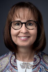Prof. Dr. Jutta Rump Porträt einer Frau mit Brille, lächelnd, in einem bunten Blazer mit einem Baum-Anstecker, vor neutralem Hintergrund.