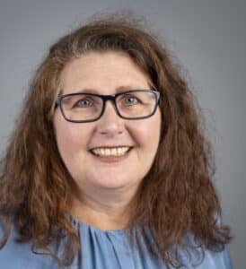Sabine Schritt Porträt einer lächelnden Frau mit lockigem, braunem Haar und Brille, vor einem grauen Hintergrund.