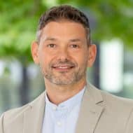 Mann mit kurzem, grauem Haar und Bart, trägt einen hellen Blazer und lächelt vor unscharfem, grünem Hintergrund.