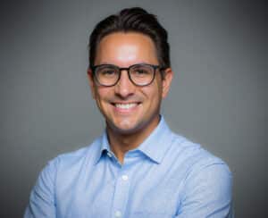 Porträt eines lächelnden Mannes mit Brille, in einem hellblauen Hemd, vor einem grauen Hintergrund.