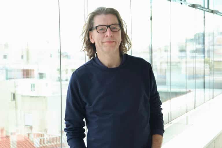 Mann mit langen Haaren und Brille steht vor großen Fenstern in einem modernen Gebäude.