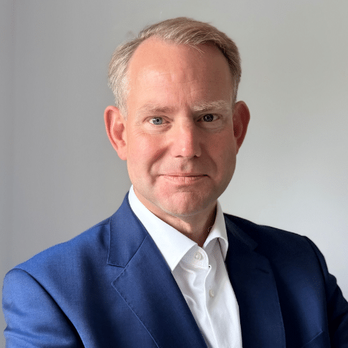 Porträt eines Mannes mit blonden Haaren, in einem blauen Anzug und weißem Hemd, vor neutralem Hintergrund.