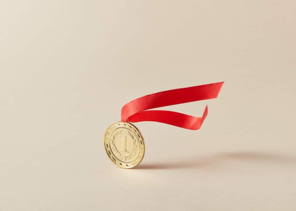 Symbolbild: Goldene Medaille mit rotem Band, die in der Luft schwebt, vor einem neutralen beigen Hintergrund. Symbol für Exzellenz, Sieg und Erfolg.
