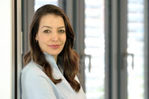 Frau mit langen, braunen Haaren in blauer Bluse, lächelt an einer Wand, im Hintergrund Fenster mit Jalousien.