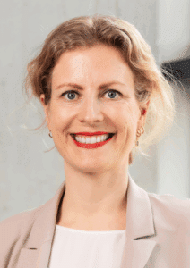 Porträt einer lächelnden Frau mit blonden, lockigen Haaren, die einen hellen Blazer trägt, vor einem neutralen Hintergr...