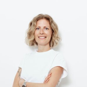 Kahle_Julia Porträt einer lächelnden Frau mit lockigem, blondem Haar, die in einem weißen T-Shirt vor einem weißen Hintergrund steht.