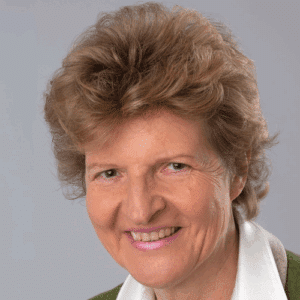 Porträt einer lächelnden Frau mit lockigem, braunem Haar und einem weißen Hemd, vor neutralem Hintergrund.