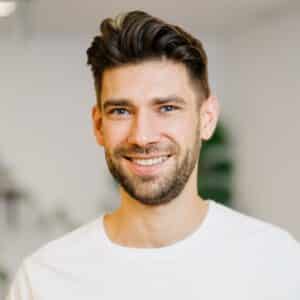 Profilbild_2024_square Lächelnder Mann mit kurzen, braunen Haaren und Bart, trägt ein weißes T-Shirt, Hintergrund unscharf.