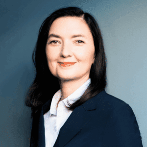 Porträt einer Frau mit langen, dunklen Haaren, die einen blauen Blazer und ein weißes Hemd trägt, vor einem blauen Hint...