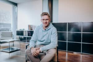 Junger Mann mit Brille in hellblauer Kapuzenjacke sitzt lächelnd auf einem Stuhl in modernem Raum mit Holzfußboden.