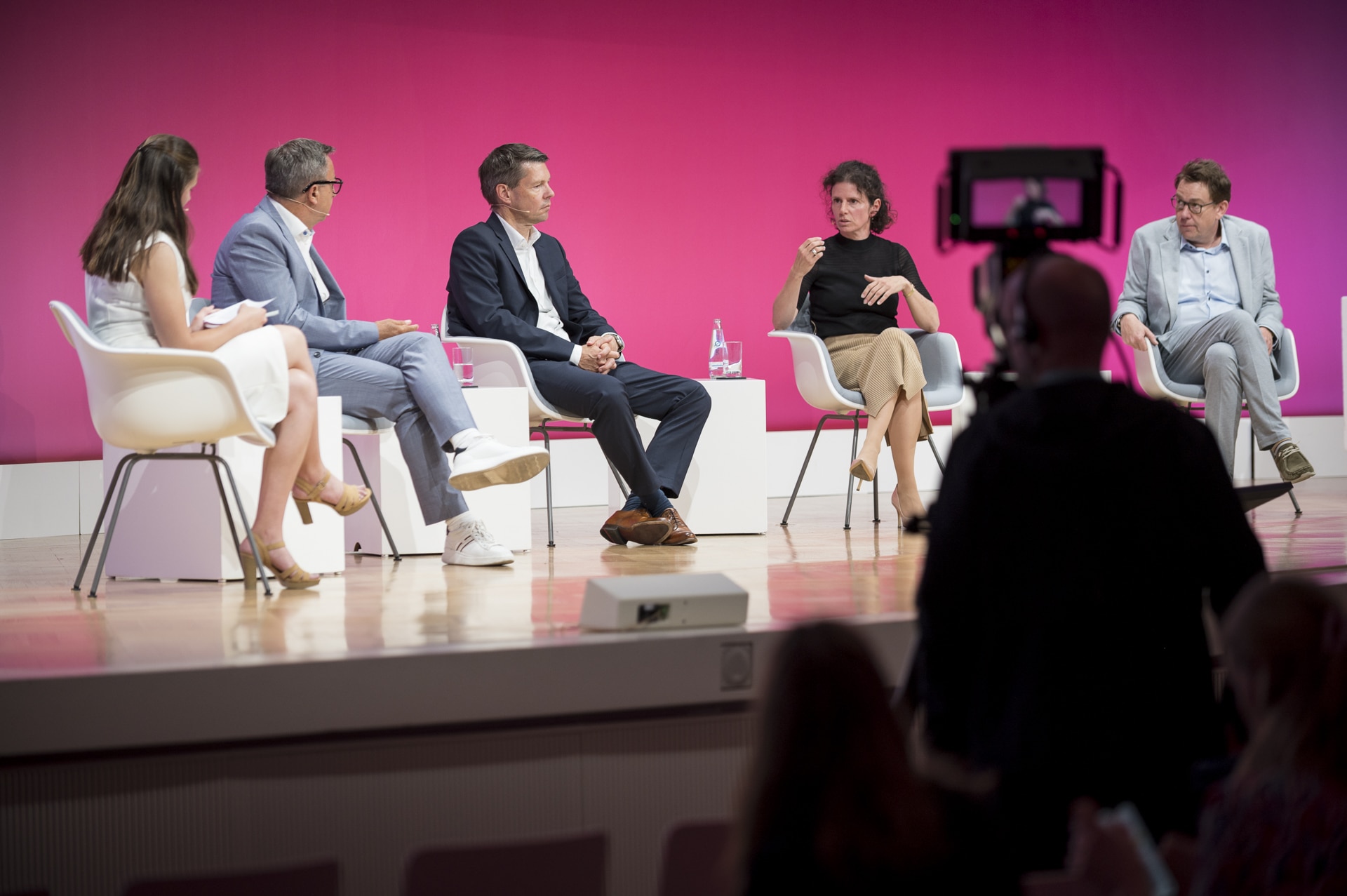 Podiumsdiskussion mit fünf Personen auf einer Bühne vor pinkem Hintergrund, einige halten Notizen.