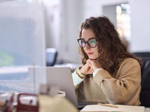 Junge Frau mit Brille sitzt am Laptop, schaut konzentriert auf den Bildschirm und denkt nach.