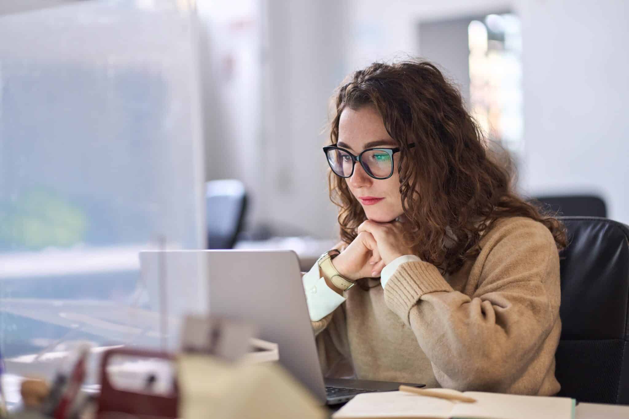 Junge Frau mit Brille sitzt am Laptop, schaut konzentriert auf den Bildschirm und denkt nach.