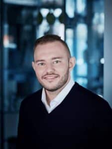 Porträt eines Mannes mit kurzem Haar, Bart und lächelndem Gesicht, in einem modernen, hellen Raum.