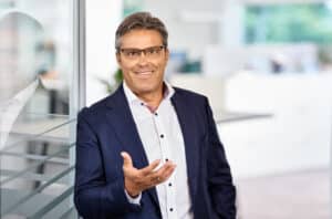 Mann in einem blauen Anzug mit Brille, lächelnd, steht an einer Glaswand und gestikuliert mit einer offenen Hand.