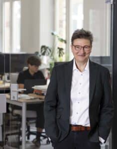 Dr. Tanja Schmidt steht in einem modernen Büro, im Hintergrund arbeitet eine Person am Schreibtisch.