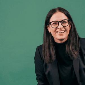 Frau mit langen, dunklen Haaren und Brille, lächelt vor grünem Hintergrund, trägt einen schwarzen Blazer.