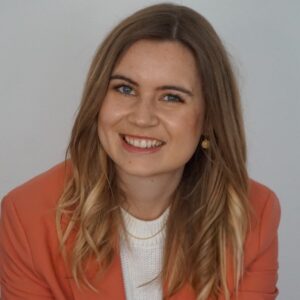 Lächelnde Frau mit langen, blonden Haaren, trägt einen rosa Blazer und sitzt vor einem hellen Hintergrund.