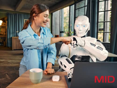 Frau mit blauen Hemd gibt einem humanoiden Roboter einen Fistbump an einem Tisch mit Laptop und Kaffeetasse.