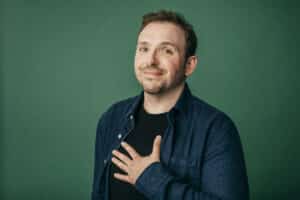Mann mit kurzem Haar und Bart, der lächelt und die Hand auf die Brust legt, vor grünem Hintergrund.