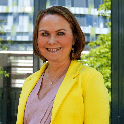 Frau mit kurzen braunen Haaren, gelber Blazer und lila Oberteil, lächelt vor einem modernen Gebäude mit grünen Pflanzen.