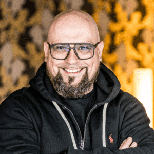 Mann mit Glatze und Bart, trägt eine schwarze Jacke, lächelt vor einem goldenen, gemusterten Hintergrund.