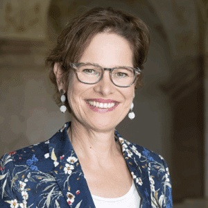 Porträt einer lächelnden Frau mit Brille, blumigem Blazer und lockigem Haar, vor einem neutralen Hintergrund.