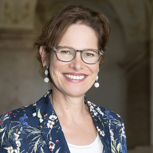 Porträt einer lächelnden Frau mit Brille, blumigem Blazer und lockigem Haar, vor einem neutralen Hintergrund.