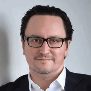 Porträt eines Mannes mit Brille, schwarzem Haar und Anzug, vor neutralem Hintergrund.