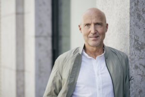 Mann mit Glatze in beiger Jacke und weißem Hemd lehnt an einer Wand, lächelt freundlich in die Kamera.