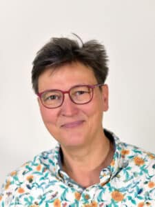 Porträt einer lächelnden Frau mit kurzen, grauen Haaren und Brille, trägt ein blumiges Hemd vor neutralem Hintergrund.