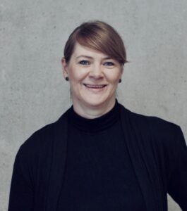 Porträt einer lächelnden Frau mit kurzen, blonden Haaren, die einen schwarzen Rollkragenpullover trägt, vor einer graue...