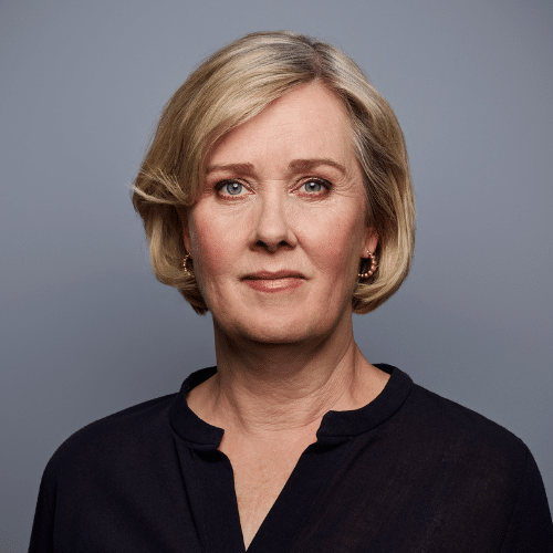 Porträt einer Frau mit blonden, schulterlangen Haaren, die in einem dunklen Oberteil vor einem grauen Hintergrund steht.