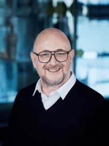 Porträt eines lächelnden Mannes mit Glatze und Brille, trägt einen schwarzen Pullover über einem weißen Hemd.