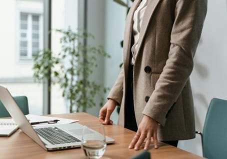 Frau in einem Anzug steht an einem Tisch mit Laptop und Notizen in einem modernen Büro. Pflanzen im Hintergrund.