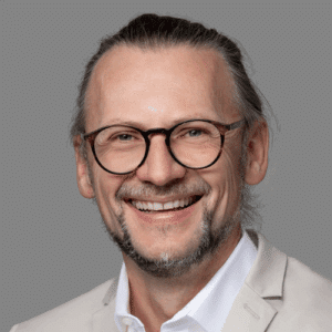 Porträt eines lächelnden Mannes mit Brille, langen Haaren und einem hellen Anzug vor grauem Hintergrund.