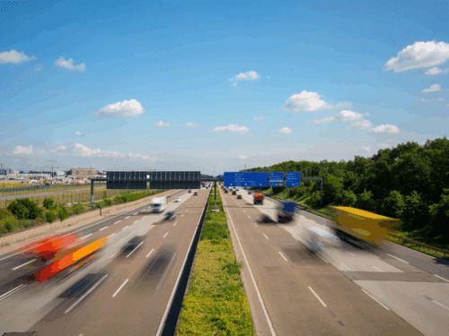 Zwei Fahrspuren auf einer Autobahn mit schnell fahrenden Fahrzeugen und blauen Verkehrsschildern unter einem klaren Himmel.