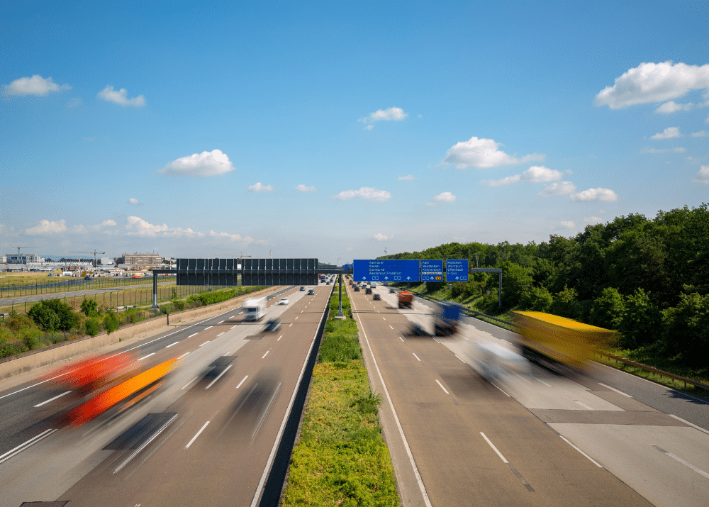 Zwei Fahrspuren auf einer Autobahn mit schnell fahrenden Fahrzeugen und blauen Verkehrsschildern unter einem klaren Himmel.