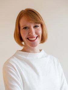 Porträt einer lächelnden Frau mit rotem Haar, die ein weißes Oberteil trägt, vor einem hellen Hintergrund.