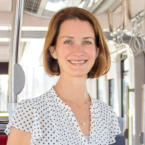 Frau mit schulterlangen, braunen Haaren und einem gepunkteten Hemd lächelt in einem modernen Bus.