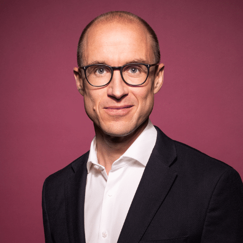 Mann mit Brille und Anzug, lächelt vor einem pinken Hintergrund.