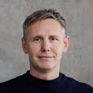 Porträt eines Mannes mit kurzem, grauem Haar, in einem schwarzen Pullover vor einer neutralen, grauen Wand.