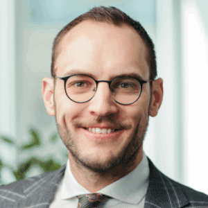 Mann mit Brille und Anzug lächelt vor einem hellen Hintergrund mit Pflanzen.