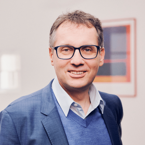 Mann mit Brille und Anzug lächelt in die Kamera, Hintergrund mit unscharfem Kunstwerk.