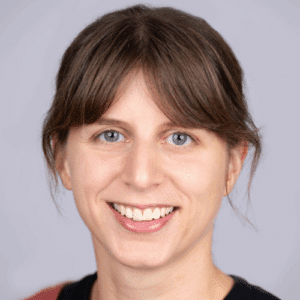 Porträt einer lächelnden Frau mit braunen Haaren und blauen Augen vor einem hellen Hintergrund.